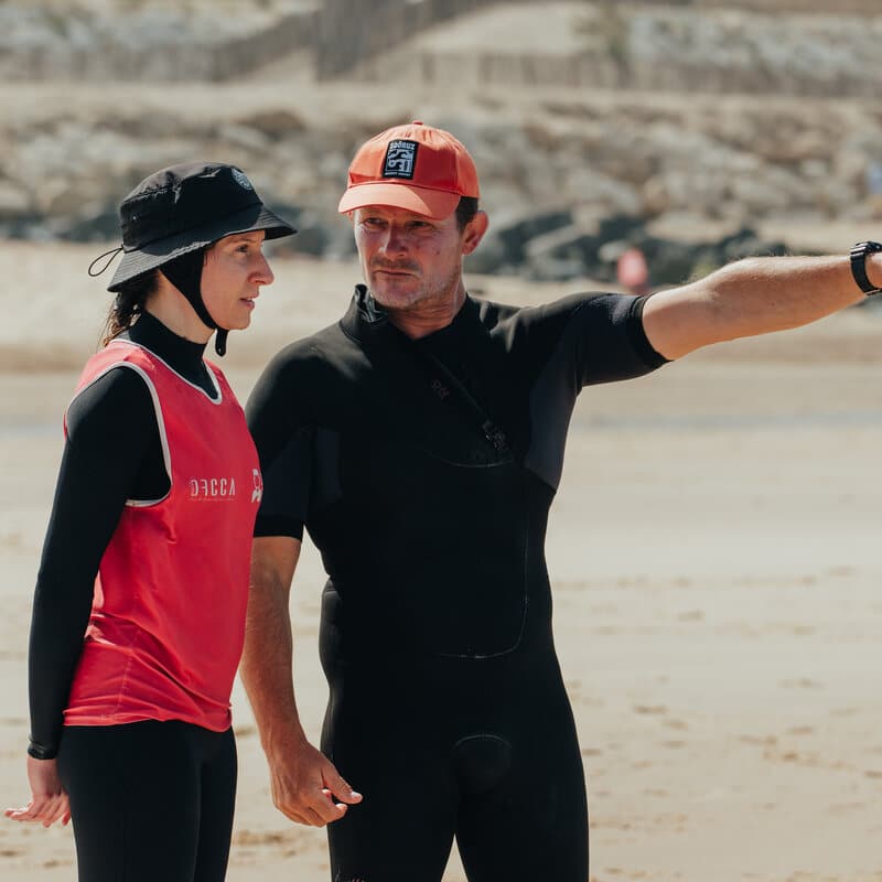 Charly Henno, en combinaison, oriente une surfeuse du bras vers un point précis de la plage avant la mise à l'eau