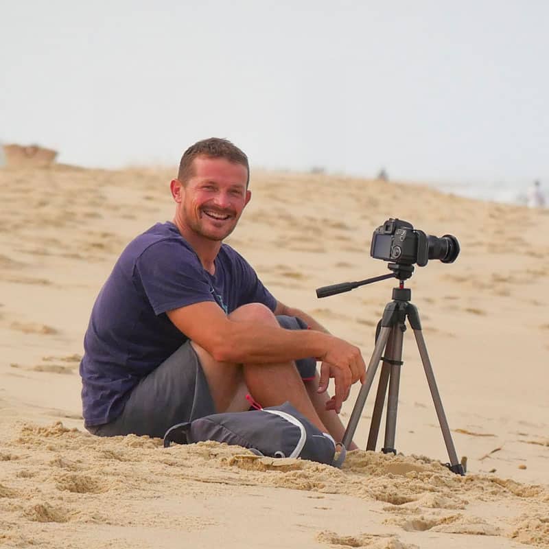 Charly Henno assis sur la plage à côté de sa caméra montée sur trépied, pendant une session de coaching