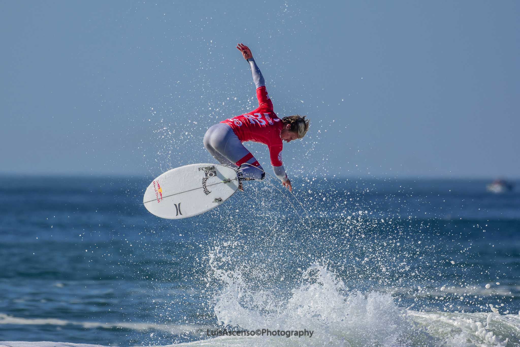 Un surfeur en lycra rouge de compétition s'envole au-dessus de la vague lors du Rip Curl Pro Portugal à Peniche, sur fond de ciel bleu