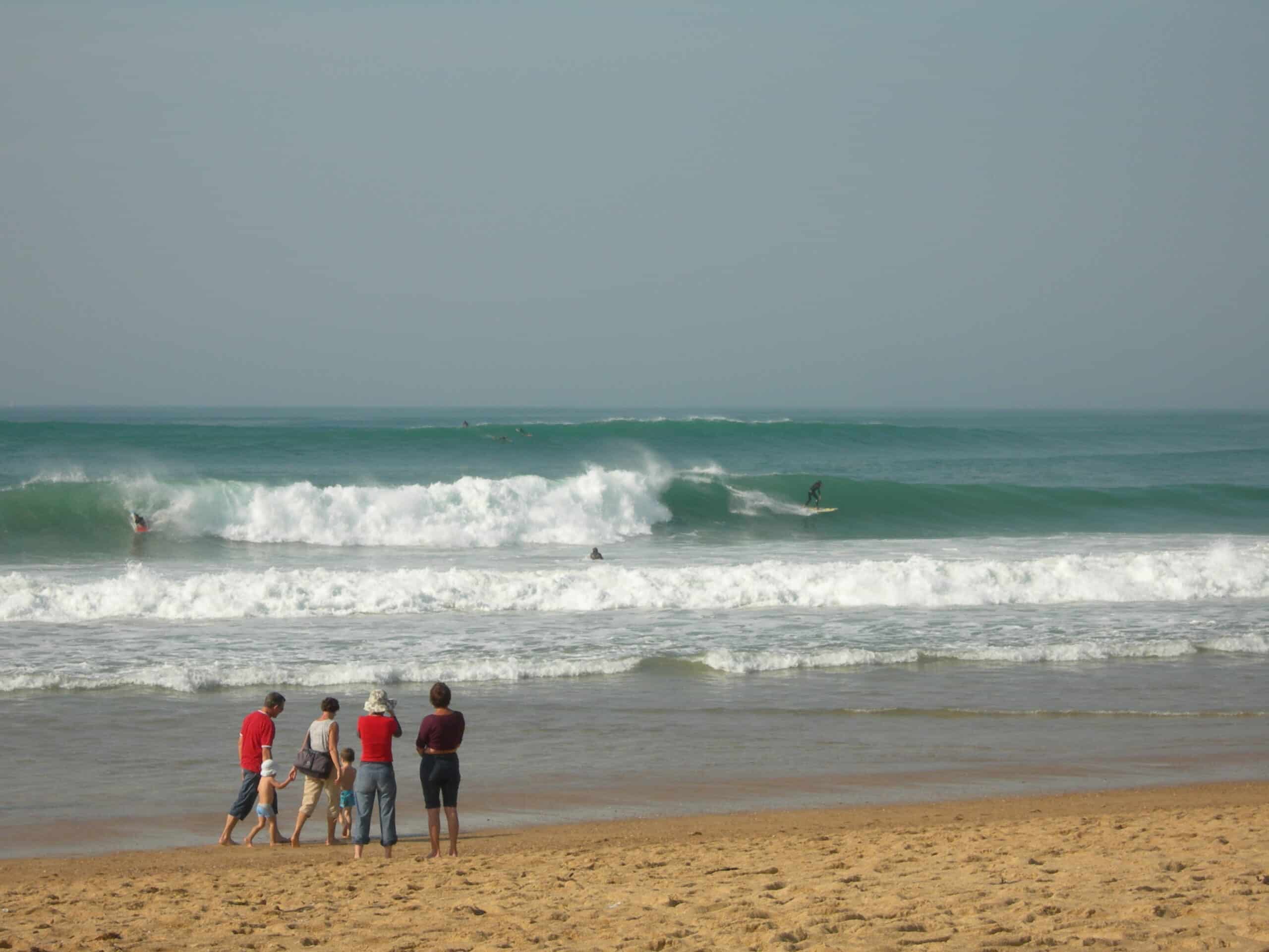 Des promeneurs regardent depuis le bord de l'eau des surfeurs prendre une vague à Hossegor