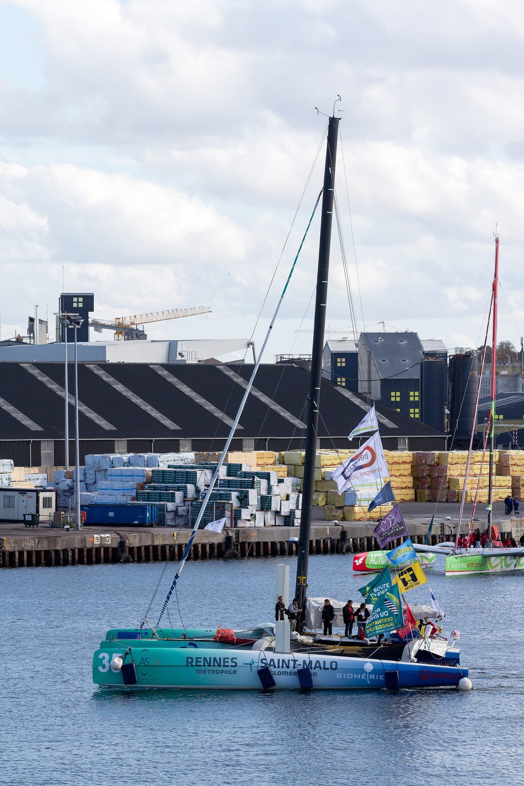 Un trimaran de 50 pieds pavoisé quitte le port de Saint-Malo, équipage et drapeaux à bord, le jour du départ de la Route du Rhum