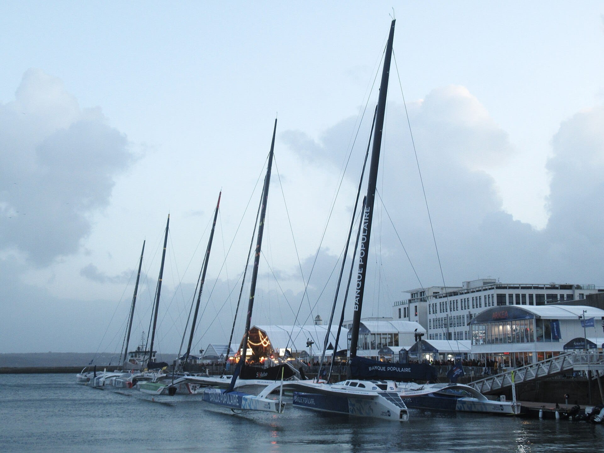 Cinq trimarans géants de la classe Ultim amarrés côte à côte au port de Brest, au crépuscule